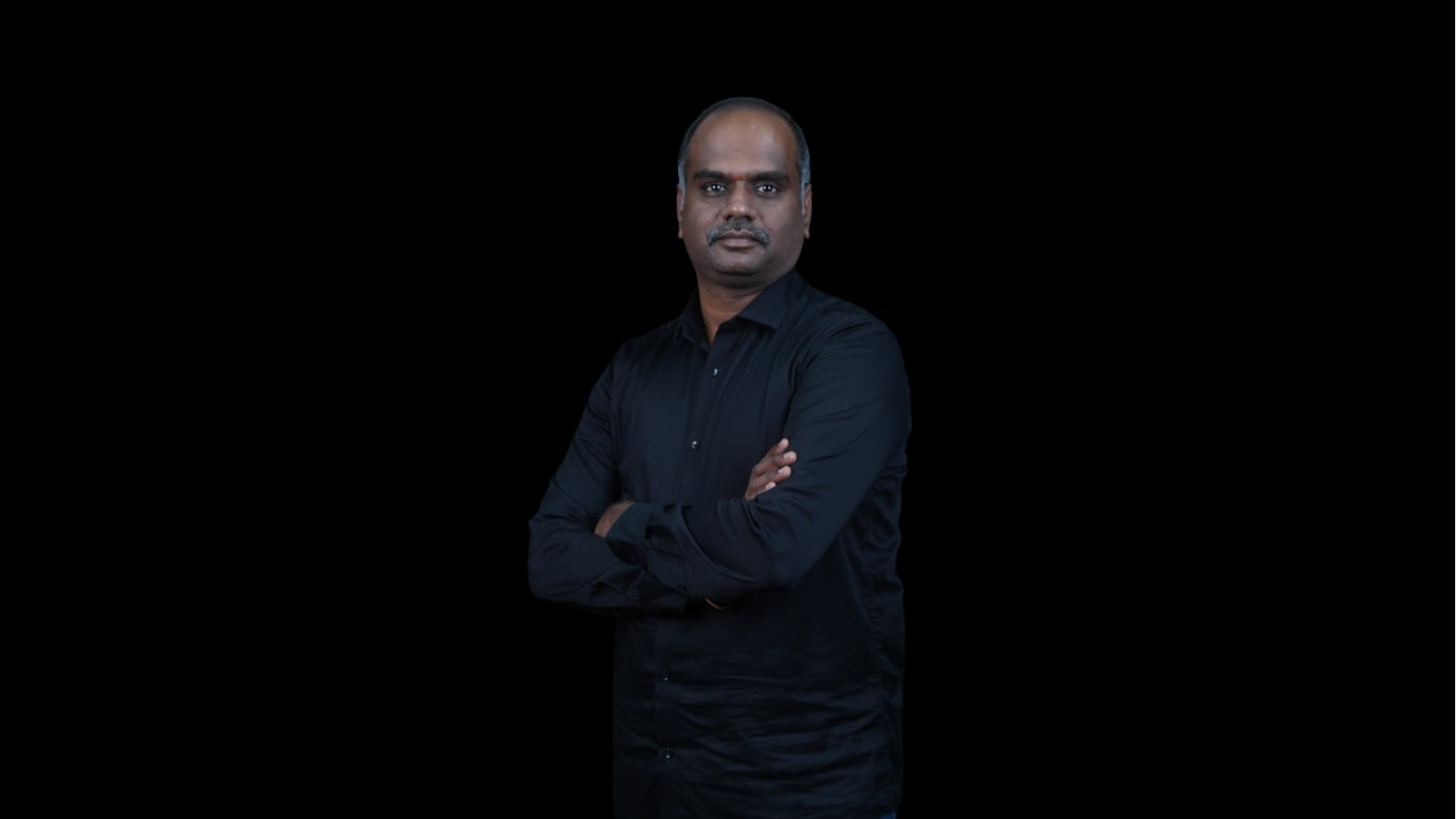 Aravind Dhandapani_BSF_IDA Professional portrait of a man in a black shirt with arms crossed against a black background.