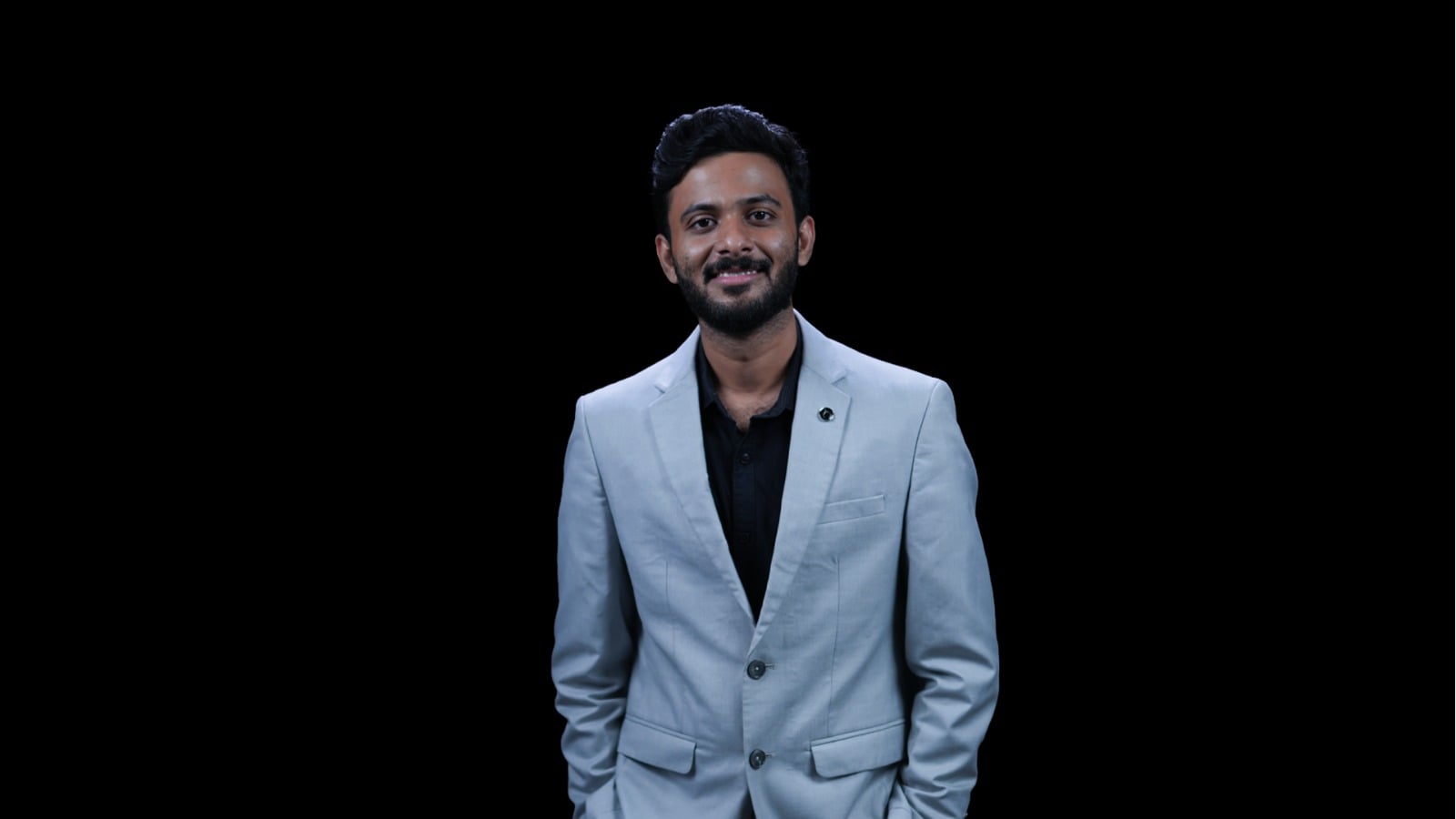 Audithyavignesh_BSF_IDA Professional portrait of a man in a light gray blazer and black shirt against a black background.