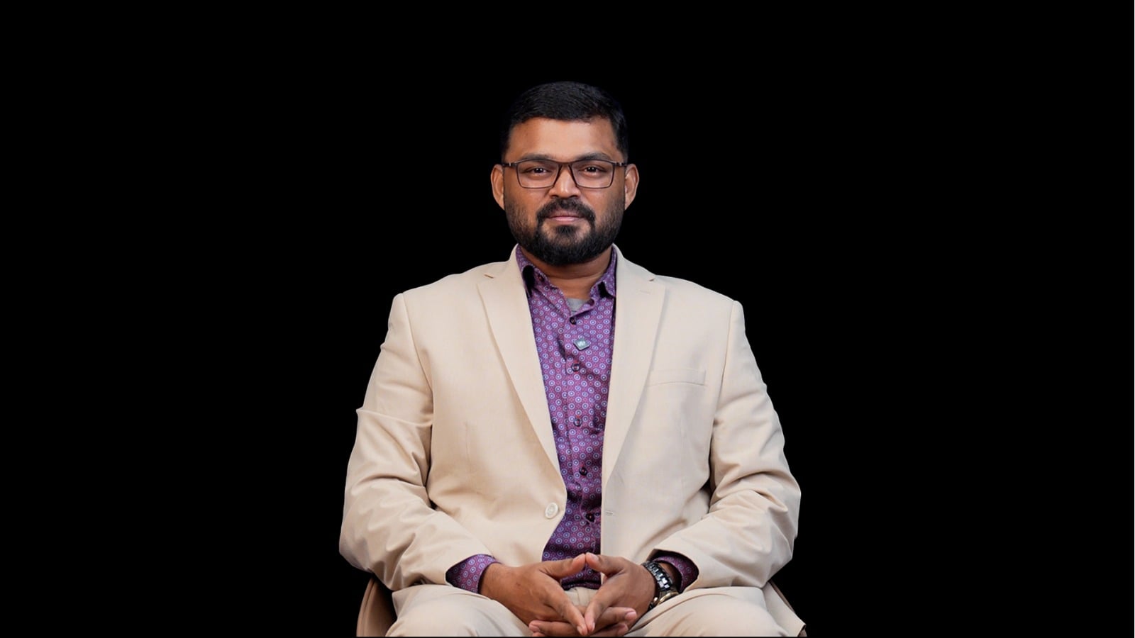 Deepak Rajaa_BSF_IDA Portrait of a man in a beige suit and purple patterned shirt, seated with hands clasped, against a black backdrop.