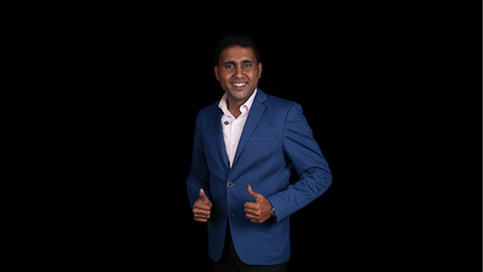 Ganesh Chandrasekaran_BSF_IDA Smiling man in a blue blazer giving thumbs up against a black background, looking at the camera.