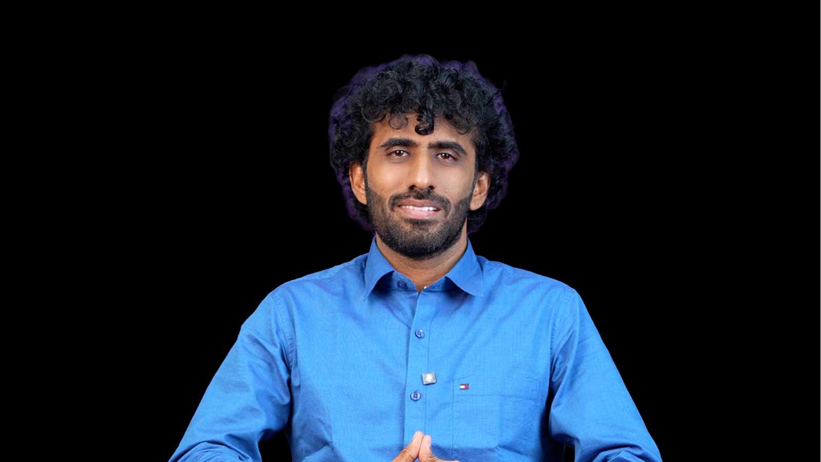 Sandeep_BSF_IDA Smiling man with curly hair and beard in a blue shirt, seated against a black background, hands pressed together in front of him.