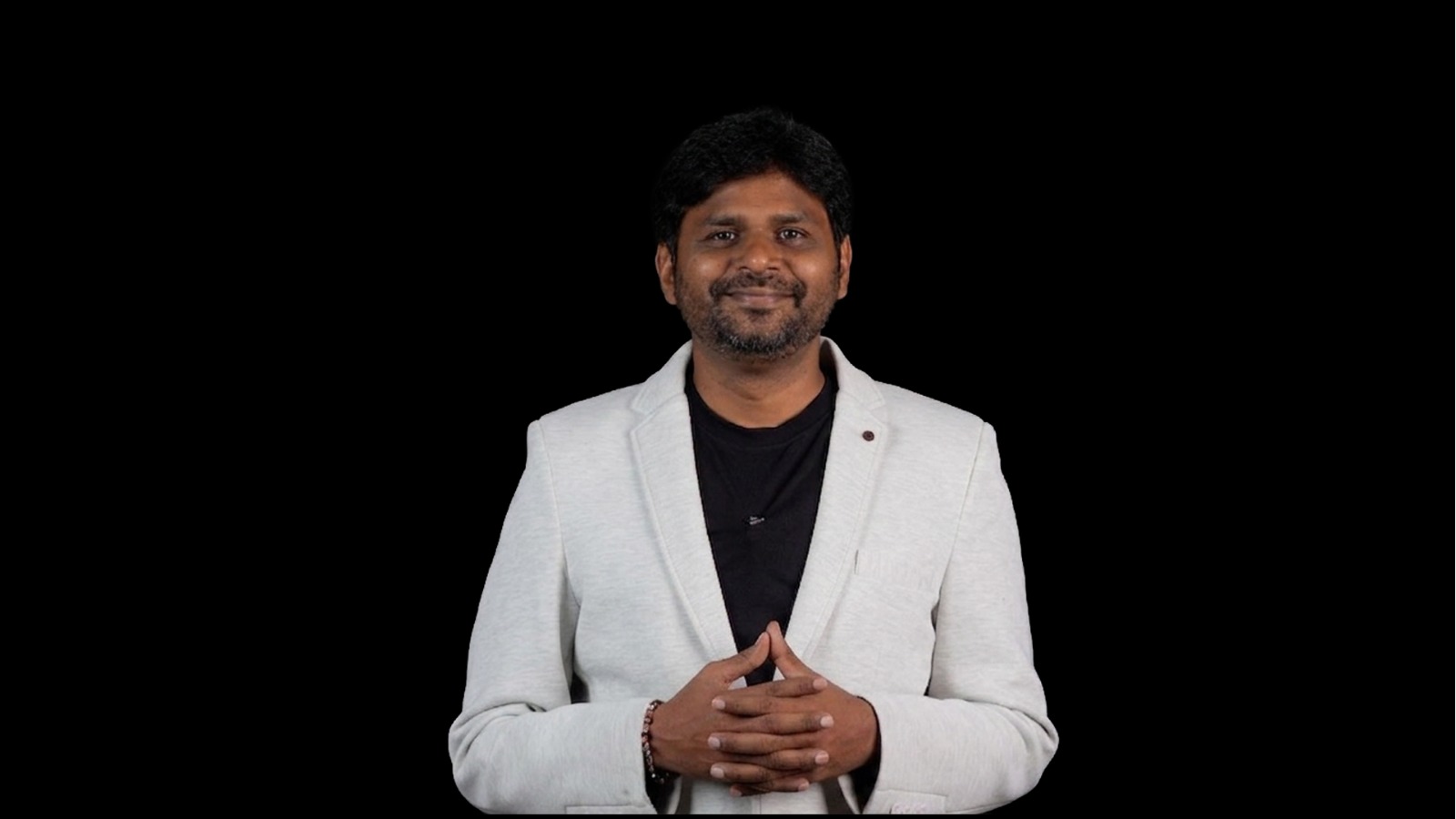 Santhosh Subramanian_BSF_IDA Portrait of a man in a light gray blazer and black shirt, hands clasped in front, smiling against a black background.